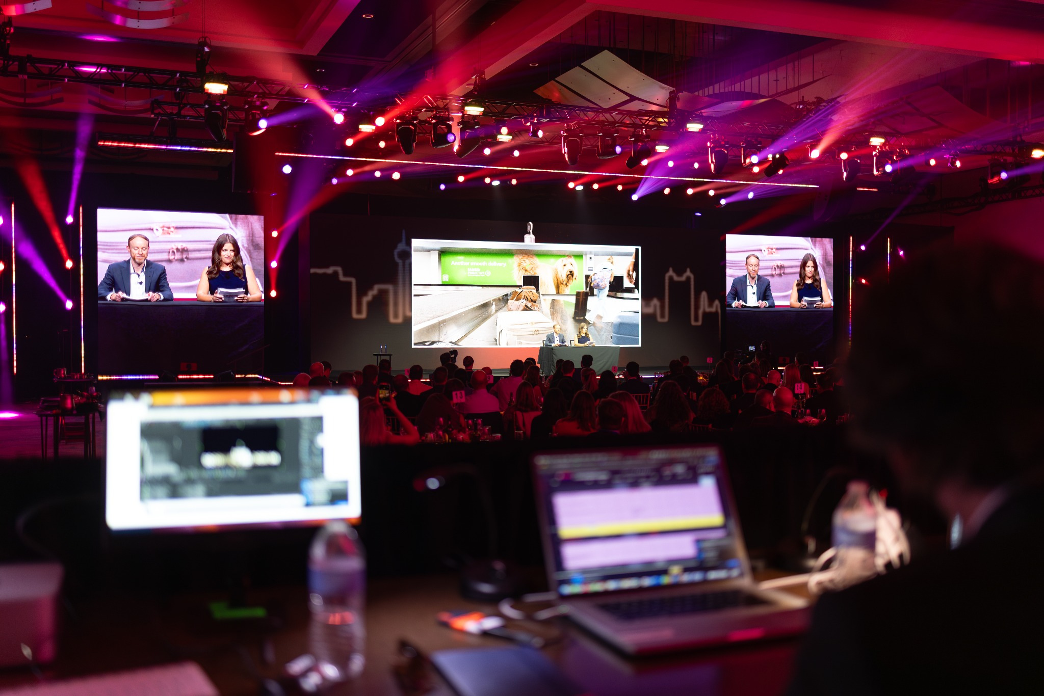 A large conference viewed from the back of the ballroom with presentation screens, stage lighting and a full audience